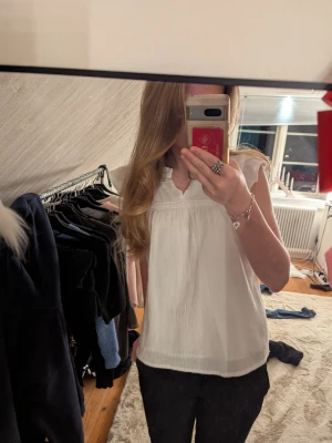 Vit blus med volang från Sansa - Supersöt vit blus från Sansa med volangdetaljer vid ärmar och hals. Blusen har v-ringning och är tillverkad i ett lätt, luftigt bomullstyg. Perfekt för en fräsch och stilren look.