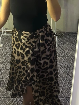 Leopardmönstrad omlottkjol - Snygg omlottkjol med leopardmönster i bruna och svarta toner. Kjolen har volangdetaljer längs kanten och knyts i midjan för en trendig look. Perfekt för dig som vill sticka ut med ett djurmönster.