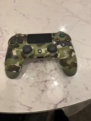 PS4 handkontroll kamouflage - Säljer en original PlayStation 4 DualShock 4 handkontroll i grön kamouflage. Kontrollen har klassisk layout med touchpad, två analoga spakar och PlayStation-knapp. Tillverkad i plast med matt finish och tydliga symbolknappar.