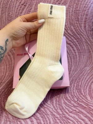 Ribbade vita strumpor från Miu Miu - Snygga ribbade strumpor från Miu Miu i en ljusvit färg. Strumporna har en klassisk, hög modell och är tillverkade i mjukt material med diskret logotyp på kanten. Perfekta för att ge din outfit en exklusiv touch.