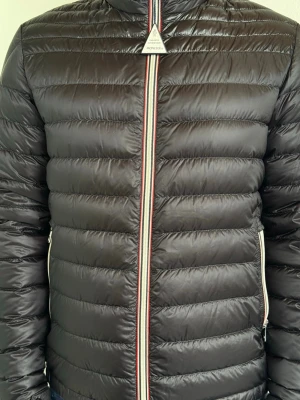 Svart dunjacka från Moncler - Snygg svart dunjacka från Moncler med quiltad design och klassisk Moncler-logga vid dragkedjan. Jackan har vita dragkedjor på fickorna och en randig detalj längs dragkedjan framtill. Perfekt för kalla vinterdagar och riktigt stilren look. Han på bilden är 175cm och väger 74kg