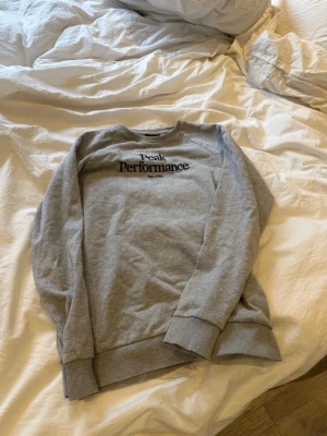 Grå sweatshirt Peak Performance - En stilren grå sweatshirt från Peak Performance med svart logotyptryck på bröstet. Tröjan har rund hals, ribbade muddar vid ärmslut och nederkant samt en avslappnad passform. 