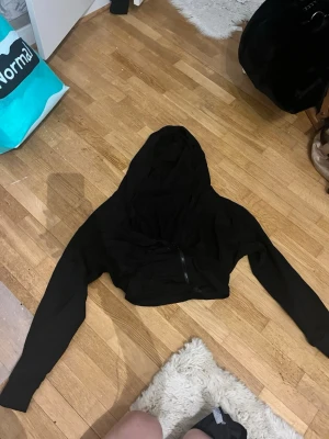 Svart croppad hoodie med dragkedja - Snygg svart croppad hoodie med dragkedja framtill och långa ärmar. Hoodien har en stor huva och är tillverkad i ett mjukt och bekvämt tyg, perfekt för en avslappnad streetstyle-look.