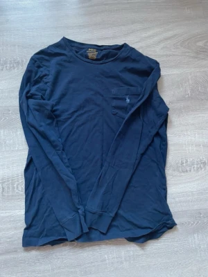 Mörkblå långärmad topp från Polo Ralph Lauren - Mörkblå långärmad topp från Polo Ralph Lauren i mjuk bomull. Klassisk rund halsringning och bröstficka med broderad logga. Perfekt för en avslappnad och stilren look.