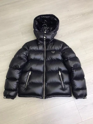 Svart pufferjacka från Prada - Säljer en svart, glansig pufferjacka från Prada med huva och dragkedja framtill. Jackan har två snedställda fickor med dragkedja och diskret logga på bröstet. Perfekt för kalla vinterdagar och riktigt trendig look.