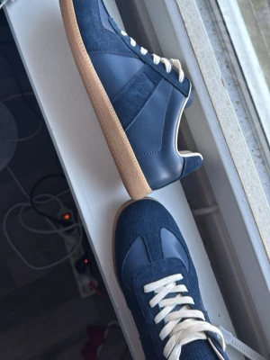 Marinblå Maison Margiela Replica - Snygga marinblå Maison Margiela Replica sneakers med ovandel i mocka och läder, vita skosnören och klassisk gummisula. Ikonisk design med retrovibbar och stilrena detaljer. Perfekta för dig som gillar minimalistisk och tidlös stil. Passar också st 43
