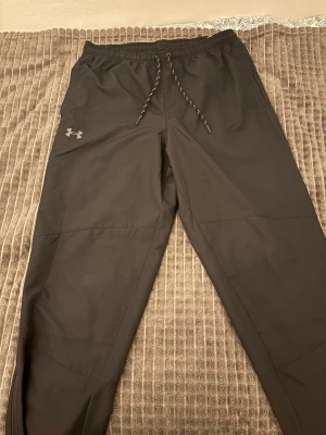 Svarta träningsbyxor från Under Armour - Svarta träningsbyxor från Under Armour med elastisk midja och snörning. Byxorna har raka ben, dragkedja vid benslut och är gjorda i ett lätt syntetmaterial som passar perfekt för sport och aktivitet. Loggan syns på vänster lår. Snygga och funktionella för gym eller löpning. Pris kan diskuteras!!!