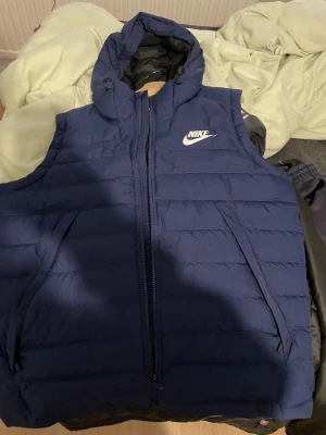 Blå dunväst med huva från Nike - Mörkblå dunväst från Nike med huva och dragkedja framtill. Västen har två snedställda fickor med dragkedja och en vit Nike-logga på bröstet. Perfekt för lager-på-lager och streetwear-stil.