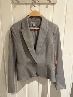 Grå kavaj från H&M - Ljusgrå kavaj från H&M med klassisk krage och enkel knäppning framtill. DS