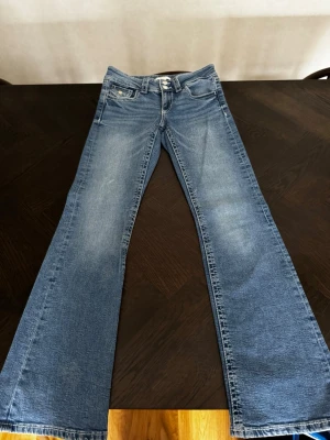 Blå bootcut jeans från Gina Tricot young  - Snygga blå bootcut jeans från Gina Tricot med låg midja och klassisk femficksdesign. Jeansen har ljusa slitningar och dubbla knappar i midjan. Perfekta för dig som gillar en retro vibe och vill ha ett par jeans med lite extra vidd nertill.