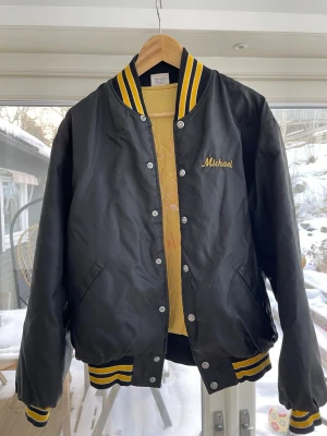 Svart bomberjacka Chestnut Ridge - Svart bomberjacka med gula och vita detaljer, broderad text 'Chestnut Ridge Razorbacks' och vildsvinsmotiv på ryggen. Namnet 'Michael' broderat på bröstet. Klassisk college-stil med ribbade muddar och krage. Litet hål på baksida av höger arm, och märker bak är lite slitet. 