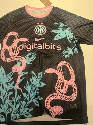 Inter Milan x Nike matchtröja svart - Unik svart Inter Milan matchtröja från Nike med rosa ormmotiv och blågröna blad. Tröjan har rund hals med rosa kant och tryckt sponsorlogga framtill. Materialet är lätt och andas, perfekt för träning eller match.