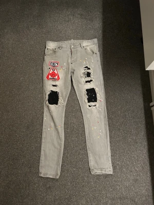 Grå ripped jeans med patchar - Säljer ett par gråa skinny jeans med stora slitningar och hål på framsidan, dekorerade med färgstänk och coola patchar som '1964' och en röd varg. Jeansen har fem fickor och är tillverkade i denim. Perfekt för dig som gillar streetwear och en unik look.