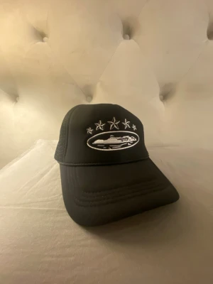 Svart truckerkeps med broderi - Säljer en svart truckerkeps med vit broderad design framtill. Kepsen har ett motiv av en bil och fem stjärnor ovanför, samt mesh på sidorna och bak för extra ventilation. Justerbar baktill för perfekt passform.