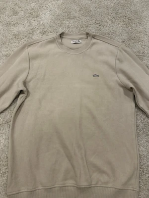 Beige sweatshirt från Lacoste - Snygg beige sweatshirt från Lacoste i slim fit-modell. Tröjan har rund halsringning, ribbade muddar och den klassiska Lacoste-loggan broderad på bröstet. Perfekt för en clean och stilren look.