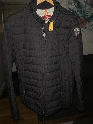 Svart dunjacka från Parajumpers - Snygg svart dunjacka från Parajumpers med quiltad design, hög krage och dragkedja framtill. Jackan har en gul detalj vid halsen och klassisk Parajumpers-logga på ärmen. Perfekt för kalla dagar och riktigt stilren look.