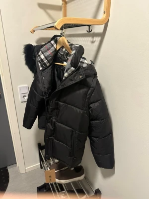 Svart pufferjacka från Burberry - Säljer en svart pufferjacka från Burberry med rutigt foder i grått, vitt och rött. Jackan har en stor huva med fuskpäls och dragkedja framtill. Märkeslogga på ärmen och praktiska fickor. Perfekt för kalla vinterdagar.