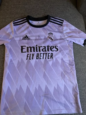 Real Madrid Benzema matchtröja lila - Säljer en lila Real Madrid matchtröja från Adidas med svart tryck, klubbmärke och sponsorlogga på bröstet. Tröjan har Benzema och nummer 9 på ryggen, svarta detaljer vid krage och axlar samt ett mönster över hela tyget. Perfekt för fotbollsfans!