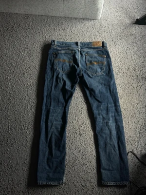 Blå jeans från Nudie Jeans - Säljer ett par klassiska blå jeans från Nudie Jeans med raka ben och femficksmodell. Jeansen har kontrastsömmar och läderpatch bak i midjan. Tillverkade i slitstark denim med snygg tvätt och diskreta slitningar. Perfekta för en avslappnad och stilren look. W31 L30