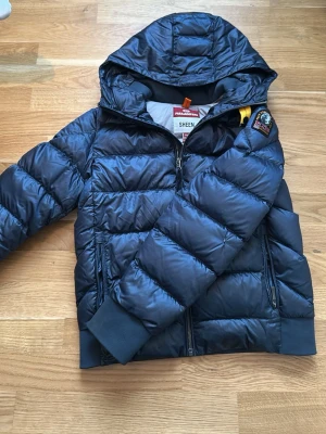 Mörkblå dunjacka från Parajumpers - Snygg mörkblå dunjacka från Parajumpers med huva, två dragkedjeförsedda fickor och ribbade muddar. Jackan har en gul detalj vid axeln och ett patchmärke på ärmen. Perfekt för kalla vinterdagar och riktigt stilren look.