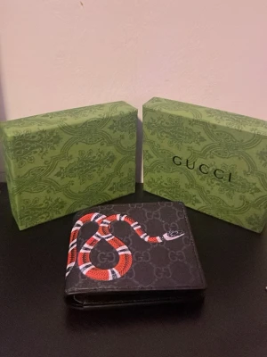 Guc4ci plånbok med ormmotiv - Snygg svart plånbok från Guc4ci med klassiskt G4-mönster och en röd, vit och svart orm tryckt på framsidan. Plånboken är i läder och har en stilren, fyrkantig form. Perfekt accessoar för dig som gillar lyxiga detaljer och unik design.