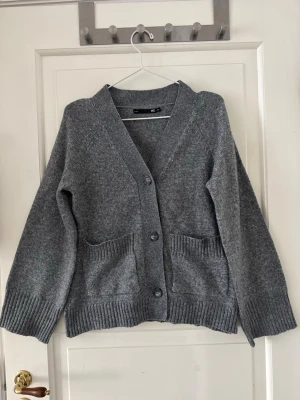 Figursydd stickadtröja - Stickad kofta ”Sabrina” från lager 157 i storlek XS men passar även S. Grå. Använd en gång och tvättad efter. Kan förekomma några noppror pga tvätt. 3% Ull och 97% polyester. Figursydd , v-ringad, vida ärmar. Skickar med en extra knapp. Inköpt för 200kr. 