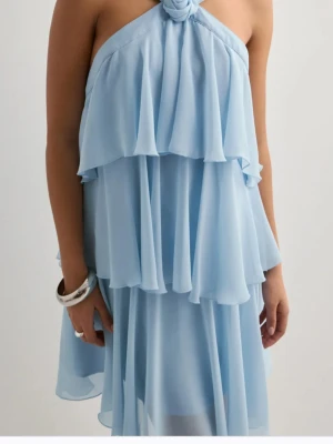 Ljusblå volangklänning med halterneck - Söt ljusblå klänning med volanger och halterneck med rosettdetalj vid halsen. Klänningen är ärmlös och har en luftig, flowy passform som ger en romantisk känsla. Perfekt för varma dagar.