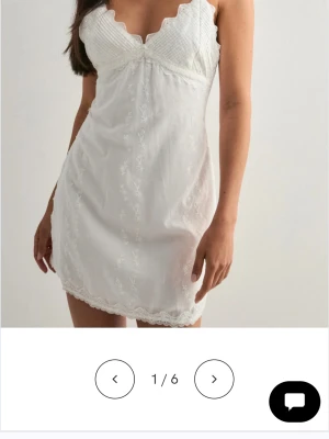 Vitt spetsnattlinne med broderier - Sött vit klänning med spetsdetaljer och broderade blommor. Modellen har smala axelband, vågig kant vid bysten och kort längd. Materialet ser ut att vara lätt och luftig.