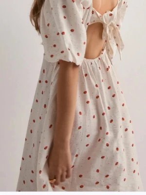 Vit klänning med röda prickar och knyt - Supersöt vit klänning med röda prickar, puffärmar och öppen rygg med knyt i ryggen. Klänningen har en luftig passform och är perfekt för varma dagar. Materialet känns lätt och andas, och volangdetaljer ger extra charm.