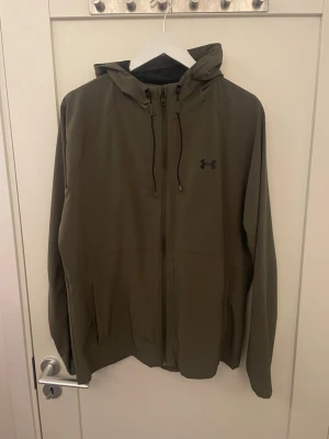 Olivgrön vindjacka från Under Armour - Snygg olivgrön vindjacka från Under Armour med huva och dragkedja framtill. Jackan har två sidofickor, justerbar huva med dragsko och diskret logga på bröstet. Tillverkad i lätt och vindtätt material, perfekt för blåsiga dagar.