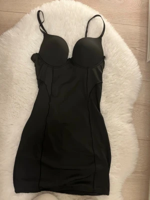 Svart shapingklänning med bygel - Säljer en svart shapingklänning med inbyggd bygel-bh och justerbara axelband. Klänningen har en figurnära passform och slätar ut siluetten. Materialet är stretchigt och bekvämt, perfekt under klänningar eller kjolar. Öppen baksida för extra komfort.