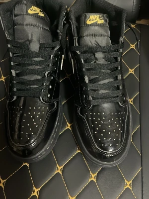 Nike Air Jordan 1 High Black/Gold - Snygga Nike Air Jordan 1 High sneakers i svart lack med gulddetaljer och svart sula. Skorna har klassisk hög siluett, svart mesh på plösen och guldfärgad Swoosh samt Air Jordan-logga på sidan. Perfekta för dig som vill sticka ut med stilrena och exklusiva detaljer.