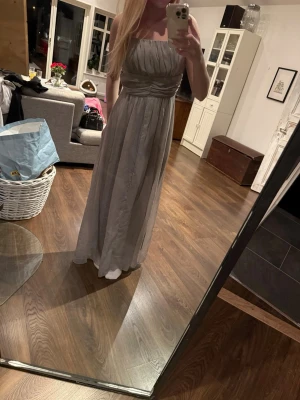 Ljusgrå  långklänning - Elegant ljusgrå långklänning med markerad midja. Klänningen har ett vackert fall och lätt rynkade detaljer upptill. Perfekt för dig som vill ha en stilren och klassisk look.