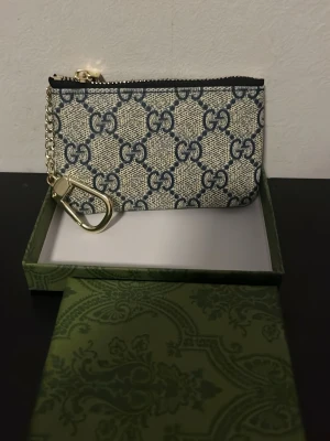 Guc4ci monogram plånbok med kedja - Snygg plånbok från Gucci i beige och blått med klassiskt G4-monogram. Plånboken har dragkedja upptill och en guldfärgad kedja med karbinhake, perfekt för nycklar eller att fästa i väskan. Tillverkad i canvasmaterial och levereras i original grön Guc4ci-box.