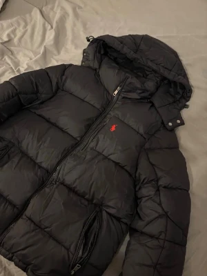 Svart dunjacka från Polo Ralph Lauren - Snygg svart dunjacka från Polo Ralph Lauren med röd broderad logga på bröstet. Jackan har huva, dragkedja framtill och två fickor med dragkedja. Perfekt för kalla vinterdagar och riktigt skön att bära. Passar även M 