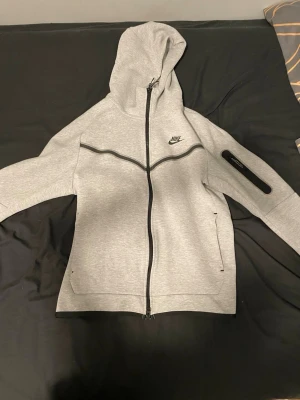 Grå Nike kit - Snygg grå hoodie från Nike med hel dragkedja framtill och svart Nike-logga på bröstet. Hoodien har huva, svarta detaljer längs bröstet och en ficka med dragkedja på ena ärmen. Tillverkad i mjukt sweatshirtmaterial, perfekt för chill eller träning.(M för män)