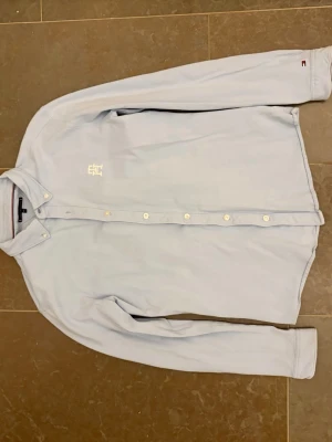 Ljusblå skjorta från Tommy Hilfiger - Snygg ljusblå skjorta från Tommy Hilfiger med klassisk krage och vita knappar. Skjortan har ett broderat vitt logomärke på bröstet och lång ärm. Perfekt för dig som gillar stilrena plagg med diskreta detaljer.