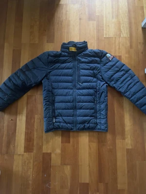 Mörkblå dunjacka från Parajumpers - Snygg mörkblå dunjacka från Parajumpers med quiltad design, hög krage och dragkedja framtill. Jackan har en bröstficka med dragkedja, två sidofickor och ett patch på ärmen. Insidan är ljusgrå och jackan är lättviktig, perfekt för kalla vinterdagar. Mörk blåig/ svart