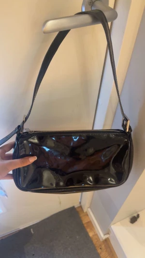 Svart lack axelväska Ginatricot  - Snygg svart axelväska i glansigt lackmaterial med rektangulär form och dragkedja upptill. Väskan har en bred axelrem och minimalistisk design utan synliga loggor eller detaljer. Perfekt för dig som gillar en stilren och trendig look.