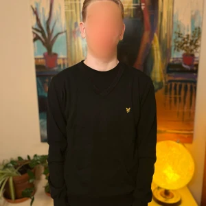 Svart V-ringad tröja från Lyle & Scott - Svart V-ringad tröja från Lyle & Scott med den klassiska gula logon broderad på bröstet. Tröjan har långa ärmar och ribbade muddar vid ärmslut och nederkant. Perfekt för en stilren och enkel look.
