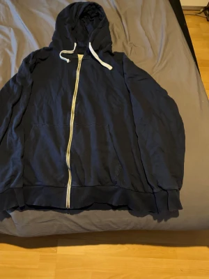 Mörkblå zip hoodie med huva - En klassisk mörkblå hoodie med dragkedja framtill och stora fickor. Tröjan har en justerbar huva med vita snören och ribbade muddar vid ärmslut och nederkant. Perfekt för en avslappnad och chill stil. Materialet känns mjukt och bekvämt.