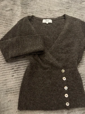 Brun omlottkofta från Gina Tricot - Mysig brun stickad omlottkofta från Gina Tricot i alpackablandning. Koftan har långa ärmar, djup v-ringning och dekorativa knappar i sidan. Perfekt för lager på lager och ger en trendig look.