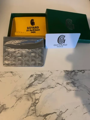 Grå kortfodral från Goy4ard - Snyggt grått kortfodral från Goy4ard med det klassiska mönstret och vit logga framtill. Tillverkad i canvas med läderdetaljer. Kommer med original ask och gul dustbag. Perfekt för kort och ID, smidig att ha i fickan eller väskan.