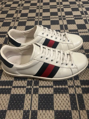 Vita Gucci sneakers med röd/blå rand - Säljer ett par klassiska vita sneakers från Gucci i läder med den ikoniska röd- och blåa sidoremsan. Skorna har vita snören, låg profil och mörkblå detalj på hälen. Perfekta för dig som vill ha en clean och stilren look med lyxig känsla.