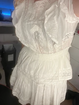 Vit kortklänning med volanger och spets - Supersöt vit kortklänning med volangdetaljer och inslag av spets. Klänningen har rund hals, korta ärmar och en öppen rygg. Tillverkad i ett lätt, luftigt tyg som passar perfekt till sommaren och studenten. Resår i midjan för en bekväm passform. Klänningen är i S, fungerar dock på mig som vanligtvis är M, kan säkert fungera på de med storlek L! Priset är som det är då jag själv köpte klänningen för 700 kr. (Klänning har lösa trådar på sidorna pga användning, däremot inget man lägger märket till)