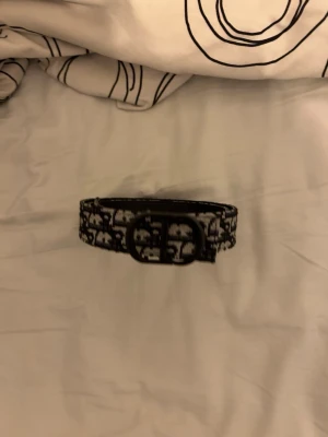 Svartvitt mönstrat bälte från Dior - Snyggt svartvitt bälte från Dior med ikoniskt mönster och dubbel D-spänne i svart metall. Bältet har en trendig design som sticker ut och passar perfekt till jeans eller kjol. Materialet är textil med tydlig Dior-logga längs hela bältet.