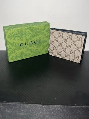 Beige G4-mönstrad plånbok från Guc4ci - Snygg beige plånbok från Gucci med det klassiska GG-mönstret i brunt. Plånboken har en stilren och kompakt design med mörka kanter och är tillverkad i skinn och canvas. Perfekt för dig som vill ha en lyxig accessoar med ikonisk logga.
