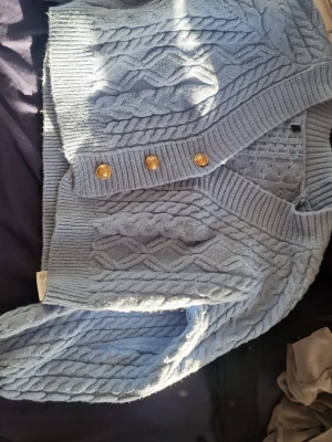 Ljusblå kabelstickad kofta med guldknappar - Superfin ljusblå kabelstickad kofta med snygga guldiga knappar framtill. Koftan har bred ribbad kant och klassiskt flätmönster. Perfekt för lager-på-lager och ger en chill men ändå uppklädd vibe. 