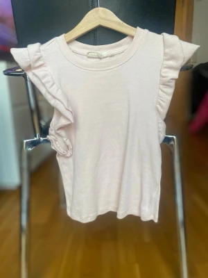 Ljusrosa volangtopp från UNIQ Studio - Supersöt ljusrosa topp från UNIQ Studio med volangdetaljer längs axlarna. Toppen är ärmlös och har en mjuk, ribbad struktur som ger en skön känsla. Perfekt att styla med jeans eller kjol för en trendig look.