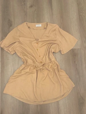 Beige blus med knappar från QUIZ - Snygg beige blus från QUIZ  med v-ringning och korta ärmar. Blusen har dekorativa guldknappar framtill och resår i midjan för en feminin siluett. Tillverkad i ett mjukt, ribbat tyg som ger en avslappnad men trendig look.